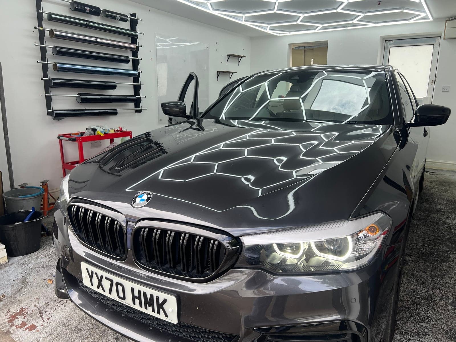 BMW 5 Series with tinted windows under hexagon lighting inside the Executive Tints & Repairs unit in Walthamstow, rolls of tint film on the wall behind
